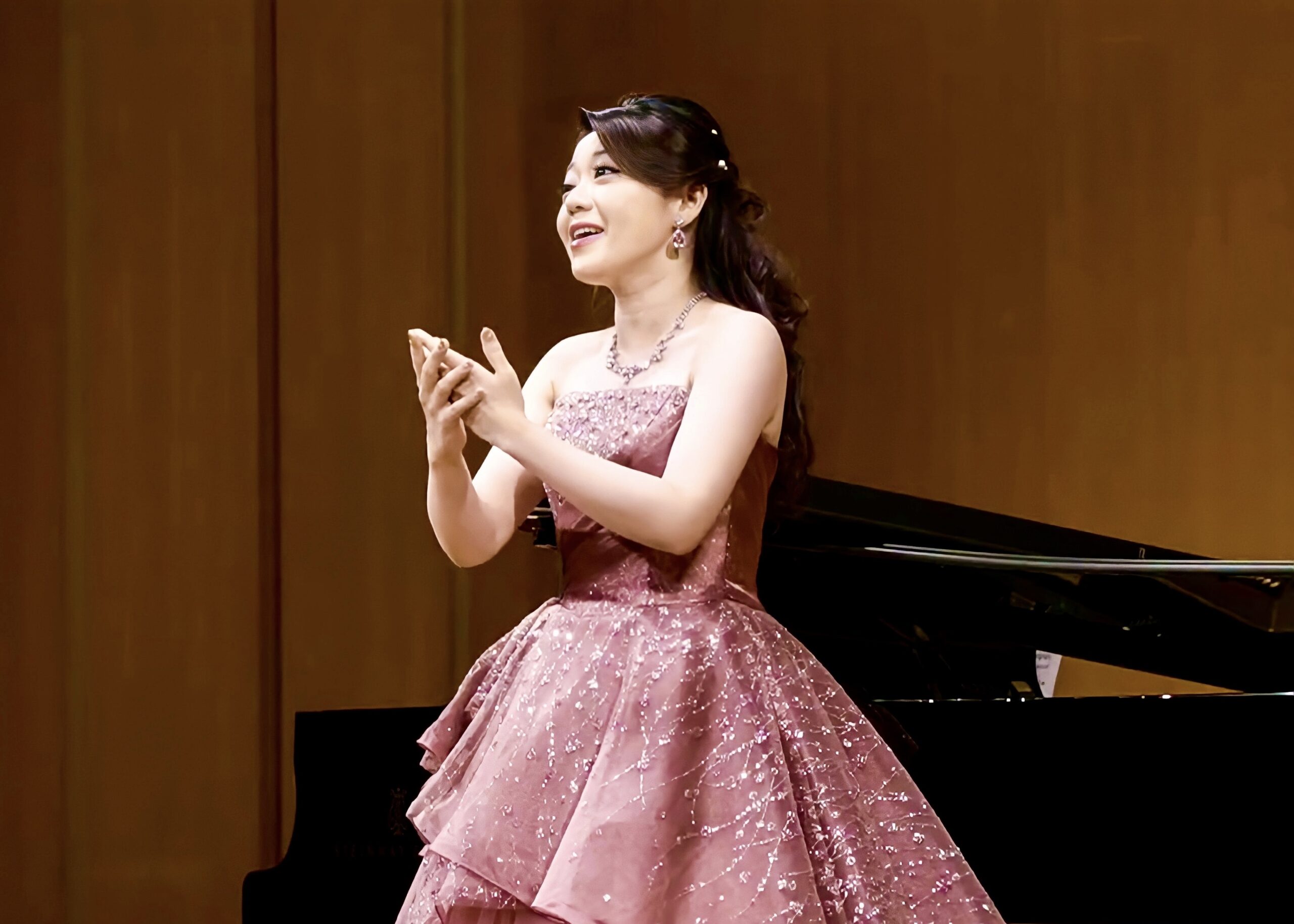 The scientist, wearing a pink gown, sings opera in front of a piano.